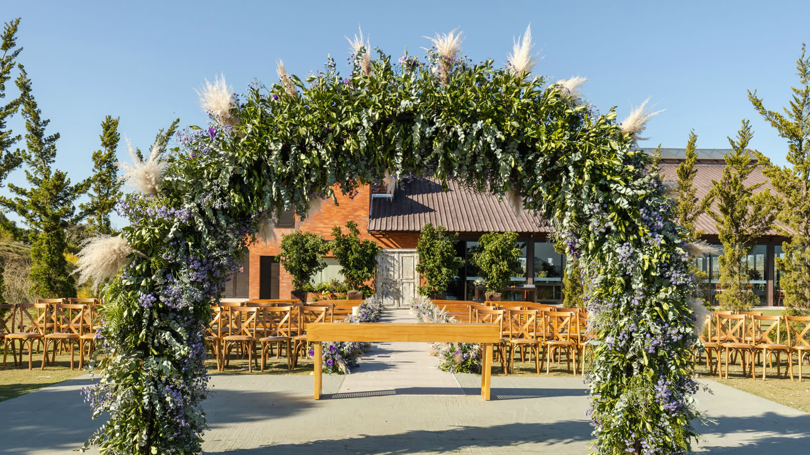 An outdoor ceremony arch of greenery, lavender and pampas grass, with rows of wooden chairs facing it